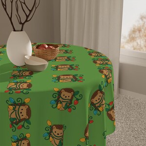 May include: A green round tablecloth with a repeating pattern of cartoon hedgehogs wrapped in Christmas lights. The hedgehogs are brown with light brown faces and are surrounded by colorful lights. A white vase, a small basket of apples, and two bowls are on the table.