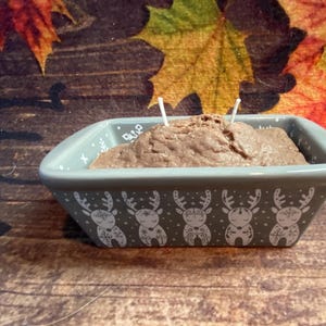 May include: A rectangular, gray ceramic dish with a brown, textured candle. The dish is decorated with white reindeer designs. Two white wicks are visible. The background features autumn leaves on a wooden surface.