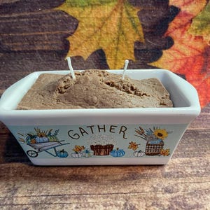 May include: A rectangular candle in a white ceramic dish with a light blue border. The candle is brown and has two white wicks. The dish has a fall-themed design with the word "GATHER".