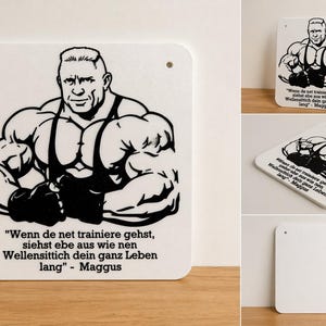 Könnte beinhalten: Weißes, rechteckiges Schild mit abgerundeten Ecken, das eine schwarz-weiße Illustration eines muskulösen Mannes zeigt. Das Schild enthält ein Zitat auf Deutsch und den Namen "Maggus". Zwei Löcher zum Aufhängen sind oben gebohrt.
