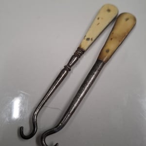 Puede incluir: Dos cuernos de zapato antiguos con mangos de marfil y ganchos de metal. Los mangos son de color amarillo pálido y tienen un aspecto desgastado y envejecido. Los ganchos de metal son de color marrón oscuro y tienen una forma ligeramente curvada.
