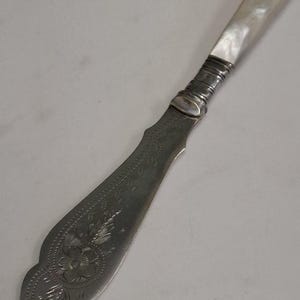 May include: A vintage butter knife with a mother-of-pearl handle and ornate floral engraving on the blade. The silver-toned knife features a decorative design and a curved blade, perfect for spreading butter or soft cheeses.