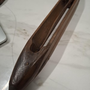 Op de afbeelding: Een donkerbruine houten weefspoel met een puntig uiteinde en een centraal kanaal. Het weefgereedschap vertoont tekenen van slijtage, wat duidt op gebruik. Het is ongeveer 25 cm lang en wordt gebruikt voor het weven van textiel.