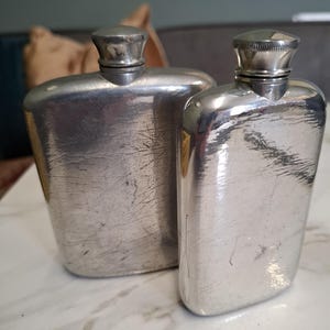 Könnte beinhalten: Zwei silberfarbene Metallflachmänner mit polierter Oberfläche und sichtbaren Kratzern. Jeder Flachmann hat einen abgerundeten rechteckigen Körper und einen Schraubverschluss. Die Flachmänner stehen nebeneinander und zeigen einen metallischen Glanz.