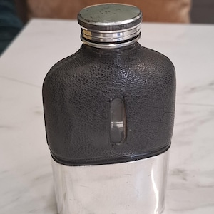 Puede incluir: Una petaca de bolsillo con una base de metal plateado y una sección superior de cuero negro texturizado. La petaca incluye una pequeña ventana de visualización vertical en el centro y una tapa de rosca plateada con un borde estriado.