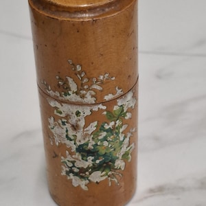 Puede incluir: Un recipiente cilíndrico de madera con tapa. El recipiente es de color marrón claro con un diseño floral en blanco y verde. La tapa es de un tono más oscuro de marrón.