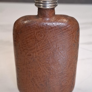 May include: A rectangular pocket flask wrapped in brown textured leather with a visible grain pattern. The flask features a rounded top and a metallic silver screw-top cap. The body of the flask has a consistent brown tone with darker brown accents.