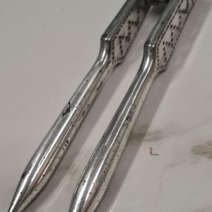 May include: A silver-toned metal nutcracker featuring two long, tapered handles with a textured, diamond-patterned grip section near the hinge. The tool has a metallic finish with visible signs of wear, including small scratches and surface scuffs.