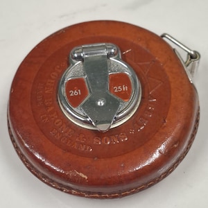 Puede incluir: Un flexómetro vintage de cuero marrón con un cierre metálico. El flexómetro tiene un centro de metal plateado con detalles rojos y el texto "25 ft" y "261". El texto "JOHN RABONE & SONS" y "MADE IN ENGLAND" está grabado en el cuero.