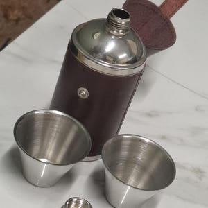 Puede incluir: Una petaca de acero inoxidable con una envoltura de cuero marrón y dos vasos de chupito de acero inoxidable. La petaca tiene una tapa plateada y una correa de cuero marrón. La petaca mide aproximadamente 13 cm de alto.