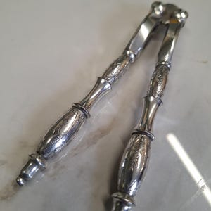 Puede incluir: Un cascanueces plateado con mangos grabados y un diseño articulado. Los mangos presentan patrones florales detallados. La herramienta está diseñada para romper nueces. El cascanueces está hecho de metal brillante.