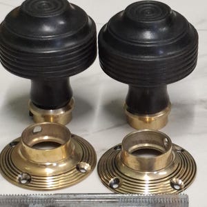 Pode incluir: Dois puxadores de porta de madeira preta com bases de latão e placas de montagem de latão correspondentes. Uma haste roscada de metal também está incluída. Os puxadores têm um topo arredondado com anéis concêntricos e uma base cilíndrica.