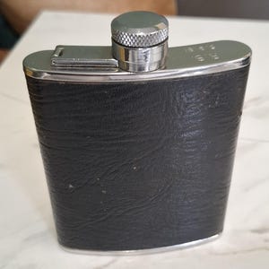 Könnte beinhalten: Ein schwarzer, mit Leder ummantelter Flachmann aus Edelstahl mit einem silberfarbenen Metallschraubverschluss. Am oberen Rand befindet sich die Gravur R.S. 5.9.81. Der Körper hat eine strukturierte Oberfläche und eine gebogene, rechteckige Form.