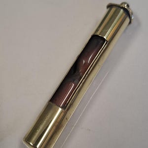 May include: A brass-colored hourglass with a dark red interior. The hourglass is cylindrical with a small cap on top. The glass section is rectangular and allows the dark red sand to flow through. The hourglass is approximately 4 inches long.