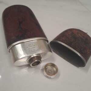 Puede incluir: Una petaca con un acabado de madera veteada en marrón oscuro y detalles en metal plateado. La petaca cuenta con una tapa extraíble y un tapón de rosca. El texto grabado en la sección plateada muestra 20, un sello y L.93.