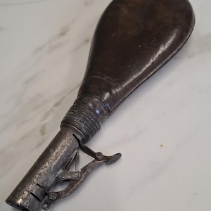 Puede incluir: Frasco de pólvora antiguo de cuero, con un cuerpo en forma de lágrima de color marrón oscuro y un pico de metal con un mecanismo de palanca. El frasco muestra signos de edad y desgaste, lo que sugiere que es un artículo vintage.
