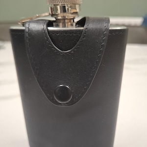 Puede incluir: Una petaca de acero inoxidable recubierta con una funda de cuero negro. La funda presenta una solapa cosida asegurada por un botón a presión negro. La parte superior tiene una tapa de rosca plateada y texturizada con un brazo metálico articulado.
