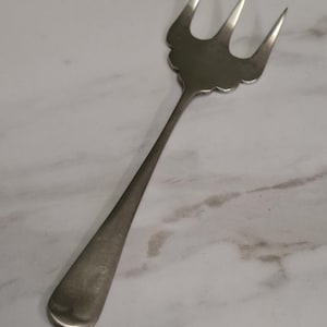 Op de afbeelding: Een zilverkleurige metalen serveervork met drie scherpe tanden en een brede, geschulpte basis waar de tanden het handvat ontmoeten. Het handvat is glad, slank en loopt taps toe naar een afgerond uiteinde, met een gepolijste, reflecterende afwerking.