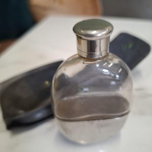 May include: A clear glass flask with a rounded body and a metallic silver base. The flask features a polished silver-toned screw-top cap. A dark, possibly leather-covered, protective case with a snap closure is positioned behind the glass flask.