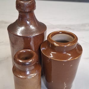 Op de afbeelding: Drie antieke bruine steengoed flessen en potten. De hoogste fles heeft een smalle hals en een afgeronde body. De andere twee zijn potten met brede openingen. Het aardewerk heeft een glanzende afwerking en een rustieke uitstraling.