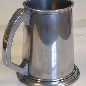 May include: A silver-coloured metal tankard with a sturdy handle and a wide, circular base. The mug has a cylindrical body and a simple, classic design. The handle is rectangular and attached to the side of the mug.