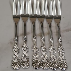 Pode incluir: Um conjunto de seis garfos de metal prateado, cada um com uma cabeça de dois dentes e um cabo decorativo em forma de caule de rosa com folhas em relevo e uma flor de rosa na base, dispostos numa fila vertical paralela para exibição.