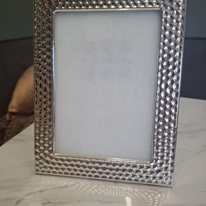 May include: A silver-colored picture frame with a textured, hammered metal design. The frame has a rectangular shape with a white interior, suitable for displaying a photograph or artwork. The frame is standing upright on a marble surface.