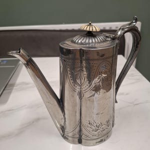 Puede incluir: Una cafetera plateada con un pico alargado y un asa curvada. La cafetera presenta intrincados diseños florales y de hojas grabados. La tapa está rematada con un adorno decorativo. La cafetera es probablemente de plata o metal plateado.