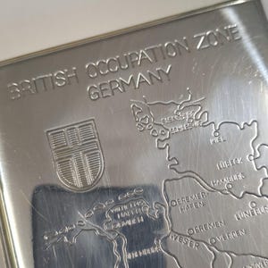 May include: A silver-colored metal item with the text "BRITISH OCCUPATION ZONE GERMANY" and a map of a region. The map includes cities like Kiel, Hamburg, and Bremen. A crest is visible on the left.