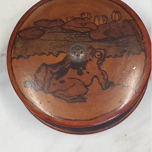 May include: Round, wooden box with a dark brown finish. The lid features a hand-painted scene of a frog and lily pads in black and brown. A small, silver metal knob is in the center of the lid.