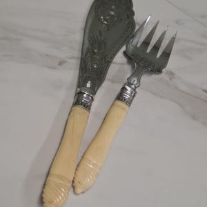 Puede incluir: Un juego de cuchillo y tenedor de servir en tono plateado con grabados florales ornamentales en las hojas y dientes de metal. Los mangos son de un material liso color crema con un diseño tallado en espiral en la base y un collar metálico.