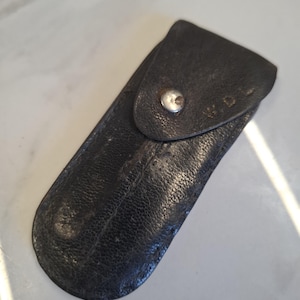May include: A black leather case with a snap closure. The case is rectangular with rounded edges and a textured surface. The initials "W.D." are visible on the flap.