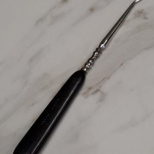 May include: A vintage crochet hook with a black handle and a silver metal hook. The handle has the text "Swiss Made" engraved on it. The hook is designed for crafting and textile work.