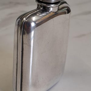 Peut inclure: Une flasque en métal argenté poli avec un corps rectangulaire légèrement incurvé et un sommet arrondi. Le récipient présente un bouchon à vis avec un bord texturé, une surface réfléchissante lisse et une couture visible sur le côté.