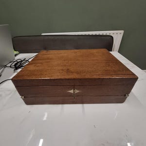 Op de afbeelding: Een rechthoekige houten doos met een donkerbruine afwerking en een glad, plat deksel. De voorzijde is voorzien van een ruitvormig metalen plaatje met een sleutelgat. Aan de linker- en rechterkant zijn kleine metalen scharnieren zichtbaar.