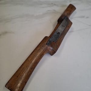 Könnte beinhalten: Ein Vintage-Holz-Hobelwerkzeug mit einer Metallklinge. Das Werkzeug besteht aus dunkelbraunem Holz und hat einen gebogenen Griff. Die Metallklinge ist rechteckig und in den Holzkörper eingesetzt. Das Werkzeug dient zum Formen von Holz.
