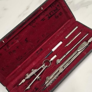 Könnte beinhalten: Ein Vintage-Zeichensatz in einem schwarzen Koffer mit rotem Samtinterieur. Das Set enthält mehrere silberfarbene Zirkel, Teiler und andere Werkzeuge. Einige handschriftliche Gleichungen sind auf der Innenseite des Koffers sichtbar.