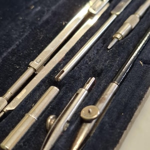 Könnte beinhalten: Ein Set aus metallenen Zeichengeräten, bestehend aus einem langen Zirkel, einem kleineren Zirkel mit Spitze und verschiedenen austauschbaren Aufsätzen. Diese Präzisionswerkzeuge haben eine metallische Oberfläche und liegen in einem dunklen Etui.