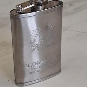 Pode incluir: Uma petaca de metal prateado com tampa de rosca articulada. A parte frontal apresenta um emblema gravado de uma criatura e o texto GLENMORANGIE SINGLE HIGHLAND MALT SCOTCH WHISKY. A petaca tem um acabamento escovado liso e formato retangular.