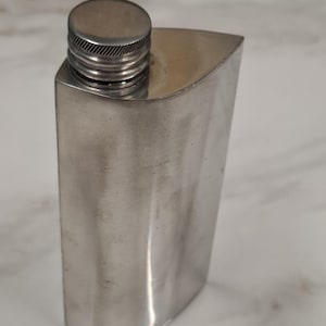 May include: A silver-coloured metal flask with a smooth, curved body and a cylindrical screw-top cap. The flask features a brushed finish and a tapered, rectangular shape with rounded edges, designed for holding liquids in a portable container.