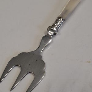 Op de afbeelding: Een vintage serveervork met een drietandige kop en een decoratief handvat. De handgreep heeft een lichtgekleurde, mogelijk benen, grip en een zilverkleurig metalen accent. De vork is gemaakt van zilverkleurig metaal en heeft een decoratieve, geschulpte rand.