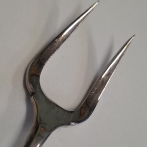 May include: Close-up of a silver, two-pronged fork. The fork has a curved design and a polished finish. The tines are sharp and pointed. The handle is smooth and rounded, with a slight curve.