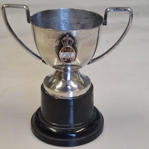 May include: A silver trophy cup with two handles and a black base. The cup features a crest with a crown and the letters "CR". The trophy is likely a vintage or antique item.