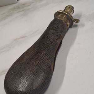 Pode incluir: Frasco de pólvora de couro antigo com parte superior de latão. O frasco tem um corpo de couro texturizado marrom escuro e um bico de latão com uma alavanca. O frasco foi projetado para conter pólvora e é um item histórico.