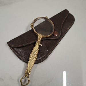 Op de afbeelding: Een vintage vergrootglas met een goudkleurige handgreep en een heldere glazen lens. De handgreep heeft een sierlijk ontwerp. Het ligt op een donkerbruine leren etui met een knoopsluiting. Het vergrootglas is een klassiek hulpmiddel.