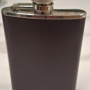 Pode incluir: Uma petisqueira retangular castanho-escura com corpo curvo e textura lisa semelhante a couro. A petisqueira apresenta uma moldura de metal prateado polido na parte superior e inferior, e uma tampa de rosca prateada com aderência texturizada.
