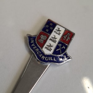 Può includere: Un segnalibro argentato con un emblema a forma di scudo. L'emblema presenta un bordo blu con la scritta "INVERCARGILL" e un disegno con una corona, stelle e altri simboli in rosso, bianco e argento. Il segnalibro è su una superficie bianca.