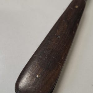 Puede incluir: Primer plano de un mango de madera para una herramienta o cuchillo. El mango es de color marrón oscuro y tiene dos remaches de latón.