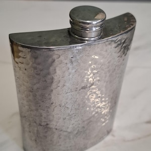 Könnte beinhalten: Ein silberfarbener Metallflachmann mit einer gehämmerten, strukturierten Oberfläche und einer abgerundeten Oberseite. Der Flachmann verfügt über einen zylindrischen Schraubverschluss mit einem kleinen, hellen Fleck auf dem Deckel.