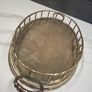May include: An oval-shaped, antique brass tray with a bamboo-style railing and handles. The tray's interior has a light brown wooden surface. The handles are curved and the railing is made of vertical bars.
